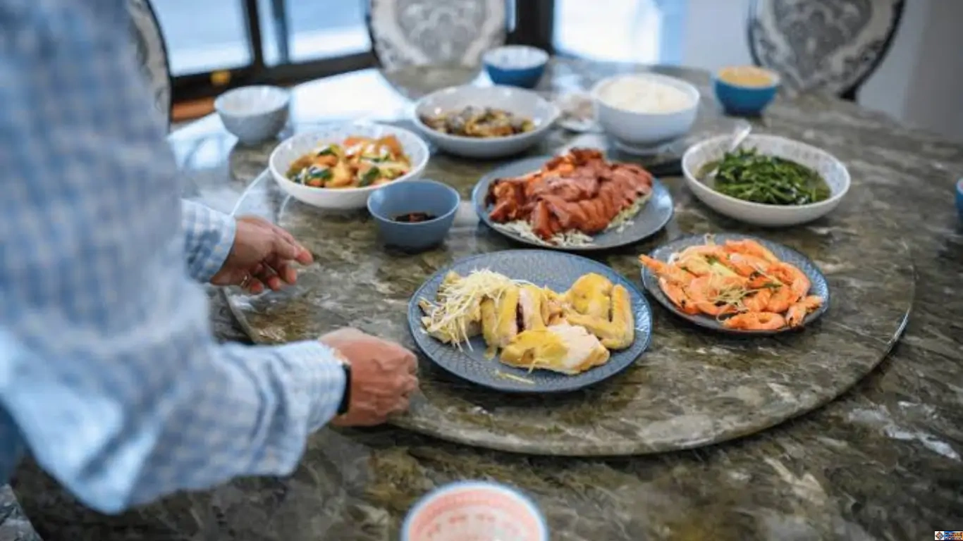 Diners reaching across a round marble table displaying the commensal architecture of shared vessels filled with family-style dishes