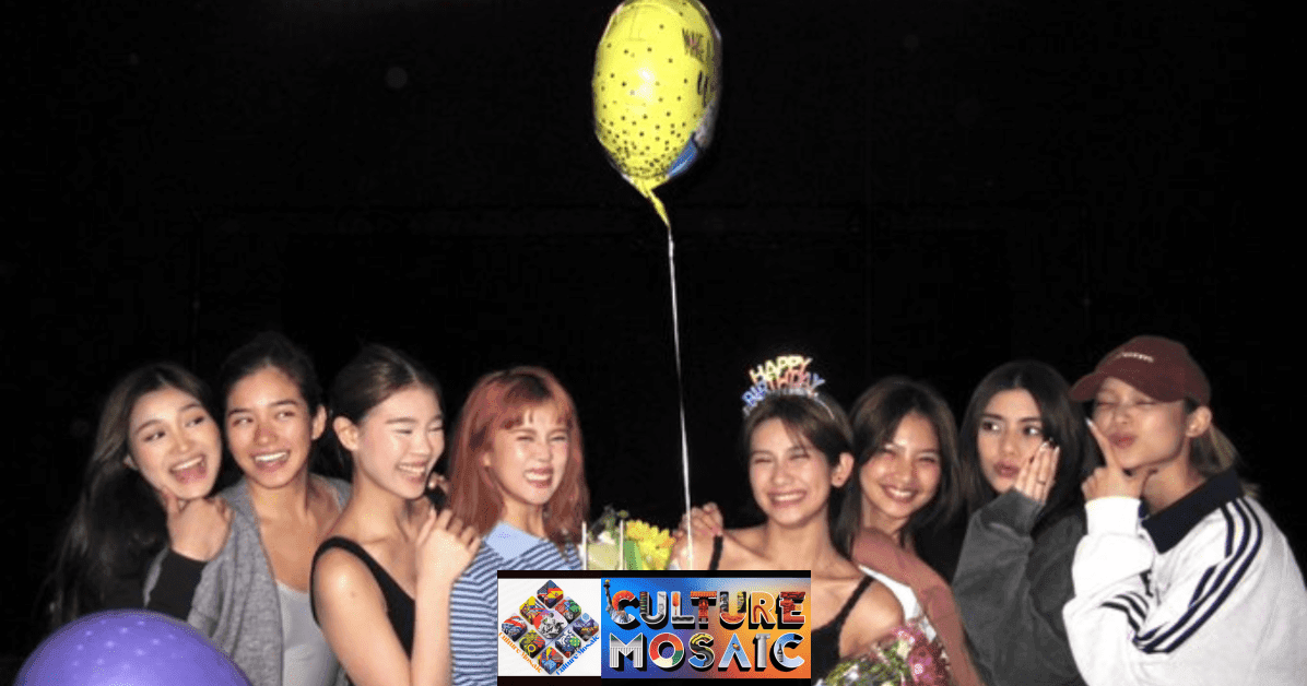 A candid indoor photo of all eight members of BINI gathered closely together around a white birthday cake with blue frosting accents. Sheena Catacutan BINI, wearing a blue striped polo, holds the cake while the members—Aiah, Jhoanna, Stacey, Maloi, Colet, Mikha, and Gwen—smile and pose playfully. A yellow star-patterned balloon floats above the center, and one member wears a "Happy Birthday" headband.