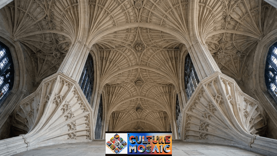 A low-angle, wide view looking straight up at the vaulted ceiling of a Gothic church. The intricate stone ceiling features fan vaulting with carved ribs spreading outward in fan shapes from clustered columns. Stained glass windows flank the sides, and carved decorative bosses adorn the central intersections of the stone arches.